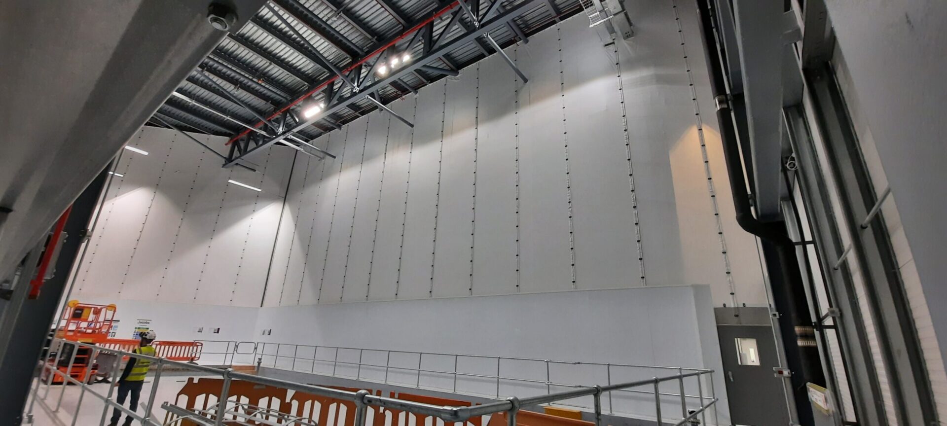 Large-scale Flexiscreen industrial curtain wall system in a pharmaceutical manufacturing facility, showing floor-to-ceiling white PVC partition panels suspended from steel roof trusses with vertical track guides, creating a clean-room grade dividing wall. A worker in high-visibility PPE and safety helmet is visible in the foreground alongside temporary scaffolding barriers and an orange scissor lift platform, with industrial LED lighting illuminating the high-bay interior.