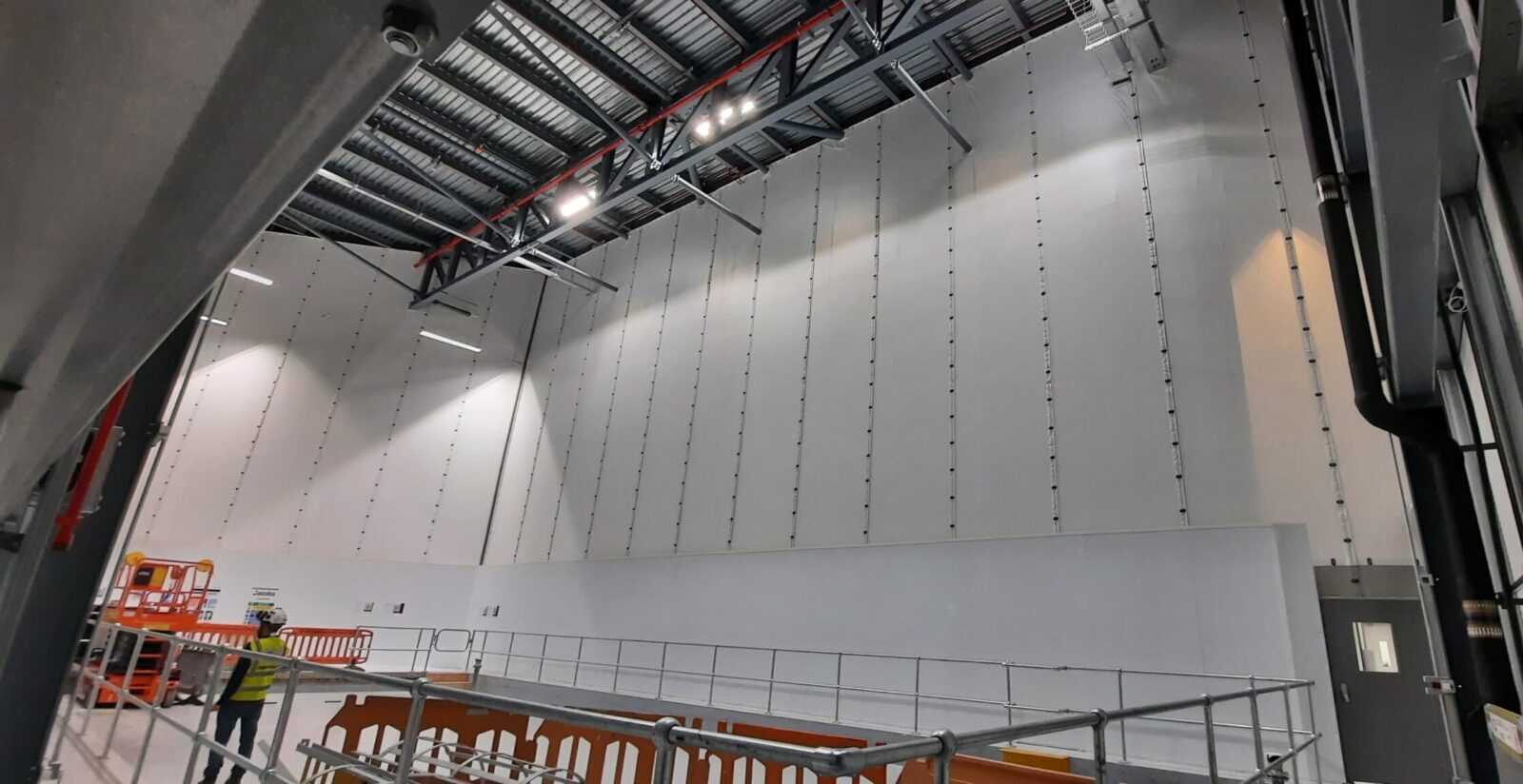Large-scale Flexiscreen industrial curtain wall system in a pharmaceutical manufacturing facility, showing floor-to-ceiling white PVC partition panels suspended from steel roof trusses with vertical track guides, creating a clean-room grade dividing wall. A worker in high-visibility PPE and safety helmet is visible in the foreground alongside temporary scaffolding barriers and an orange scissor lift platform, with industrial LED lighting illuminating the high-bay interior.